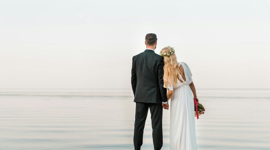 back view of wedding couple standing on beach with wedding bouquet and looking at sea